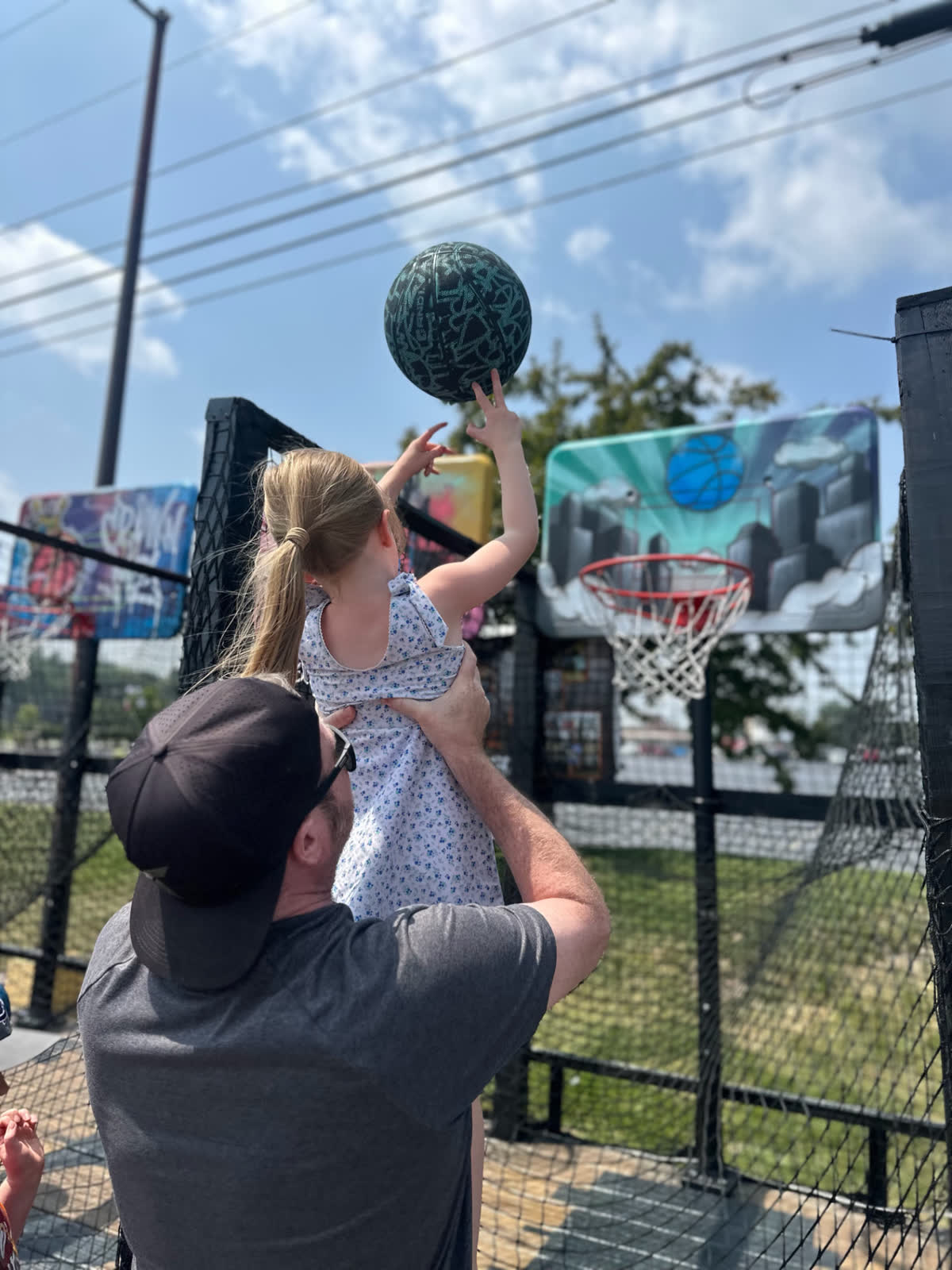 An adult lifts a young girl up so she can shoot a basketball at one of the rims.