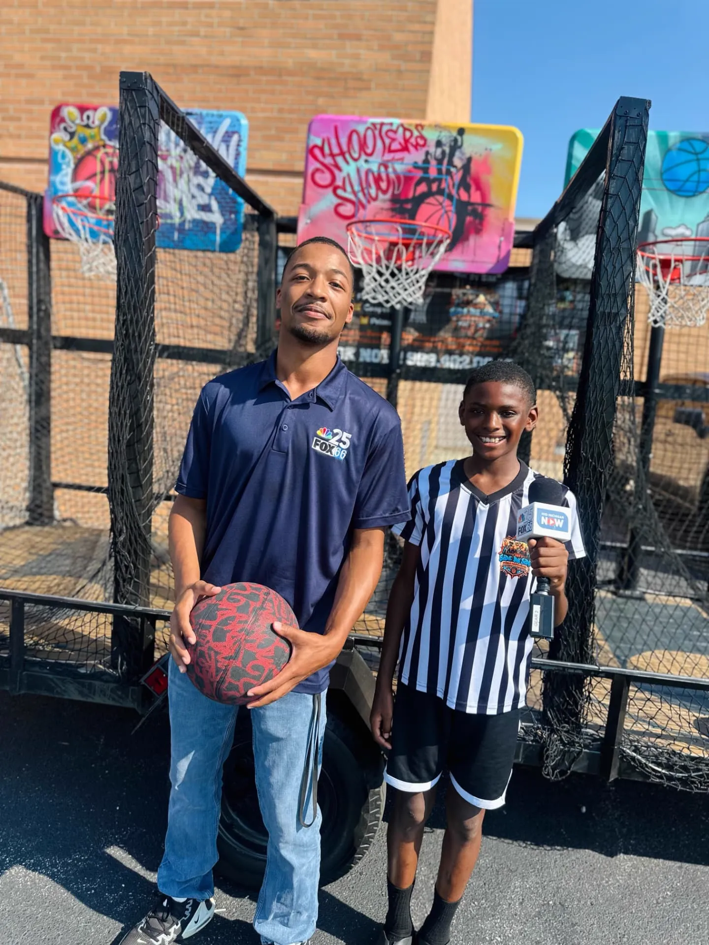 A local news reporter stands with a young Side By Side Mobile Hoops team member holding a station microphone in front of the trailer.