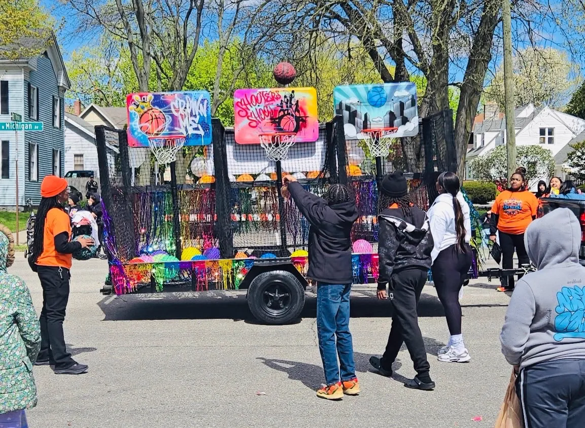 A child shoots a basketball at the Side By Side Mobile Hoops trailer during a neighborhood parade, with staff and spectators watching from the street.