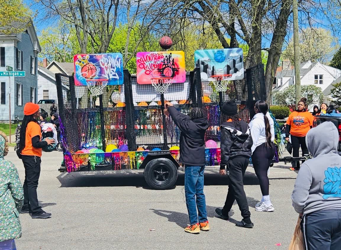 A child shoots a basketball at the Side By Side Mobile Hoops trailer during a neighborhood parade, with staff and spectators watching from the street.