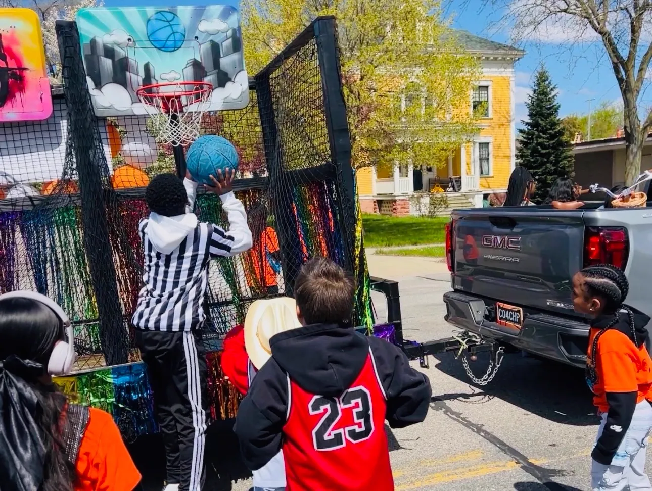 The hoops trailer hitched to a pickup truck at a parade while a child shoots at the city-skyline backboard.