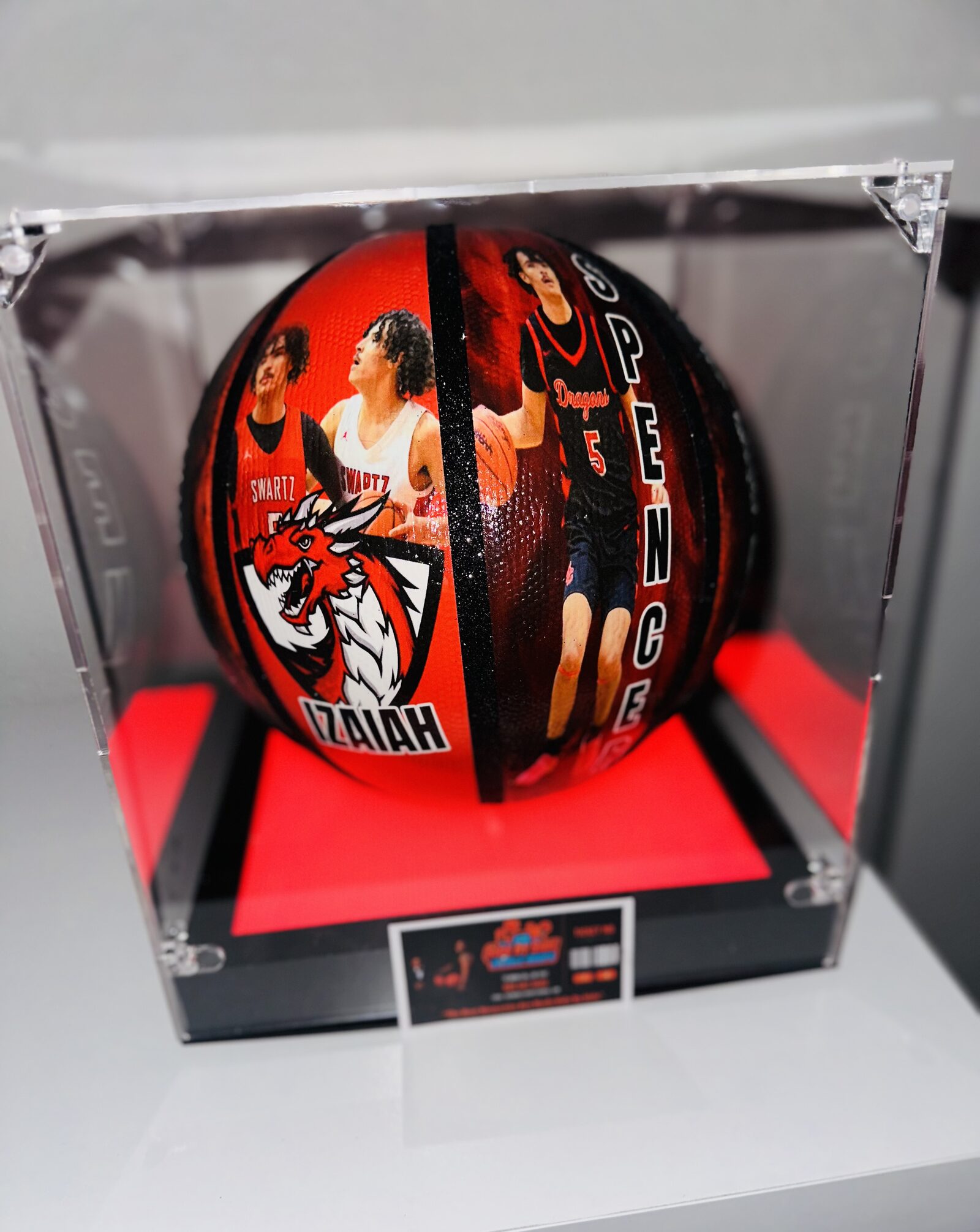 A custom full-wrap printed basketball for player Izaiah Spencer in a lit acrylic display case, showing game photos and the Swartz Creek Dragons logo.